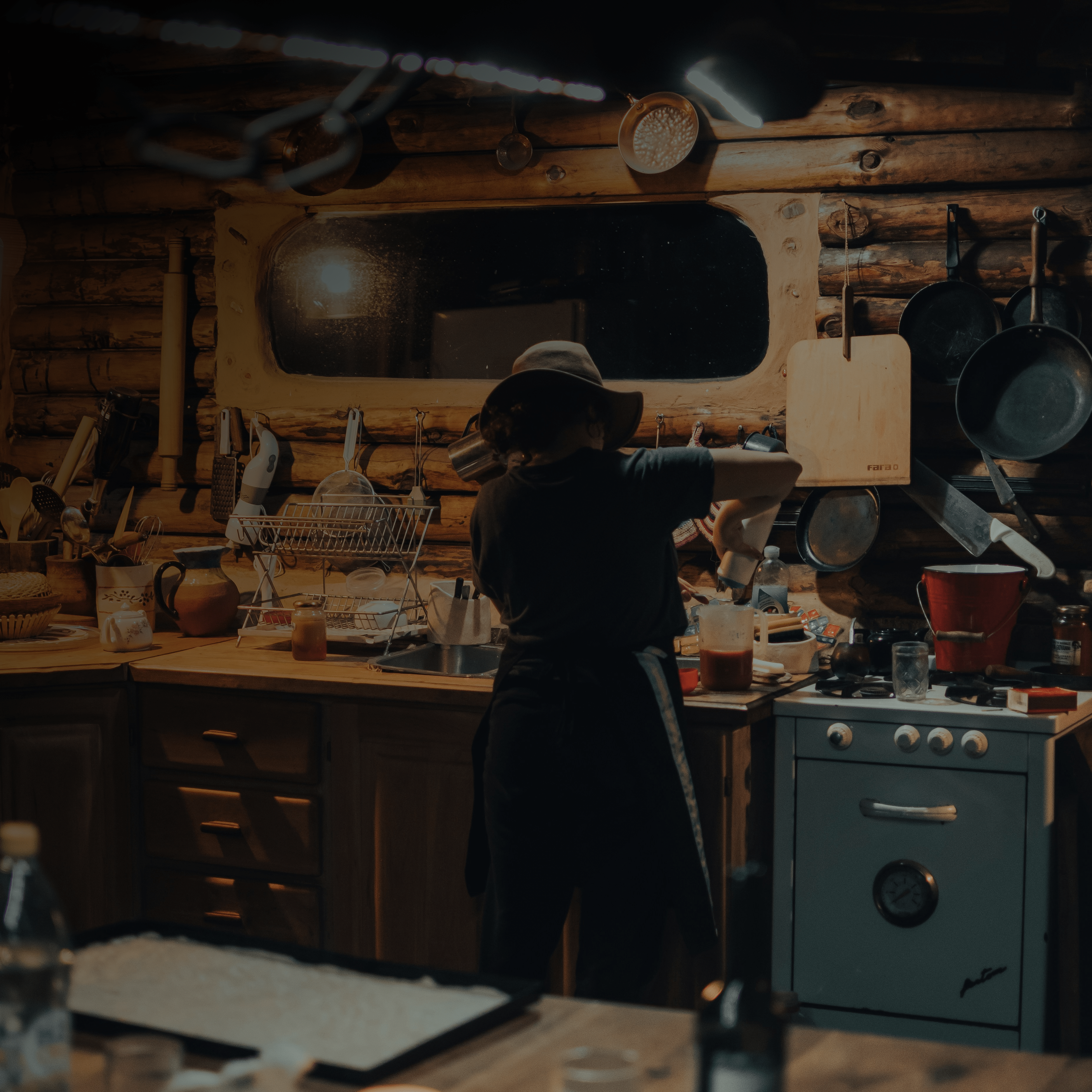 A person wearing a wide-brimmed hat and apron prepares food in a rustic, warmly lit log cabin kitchen filled with hanging pots, wooden cabinets, and vintage appliances.